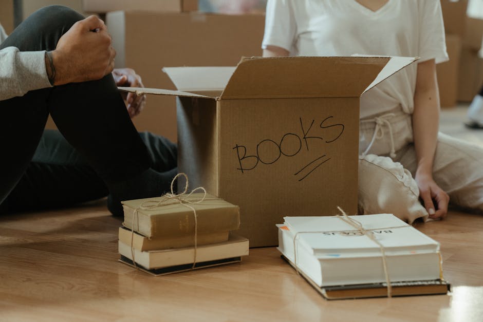 Inside a home, a person is seated on a wooden floor with their legs crossed, packing books into an open cardboard box labeled 'BOOKS' with black marker. Several stacks of books, tied with string or rubber bands, are positioned on the floor nearby, ready for transport. The individual is wearing grey and the child sitting nearby is dressed in light-colored clothing, sitting cross-legged with hands resting on their lap. In the background, other cardboard boxes and packing materials are visible, indicating an ongoing packing process during a home relocation. The scene is well-lit, emphasizing the careful organization of packed books and moving supplies, reflecting typical furniture transport and packing activities associated with residential removals, as provided by Man With a Van Finchley.