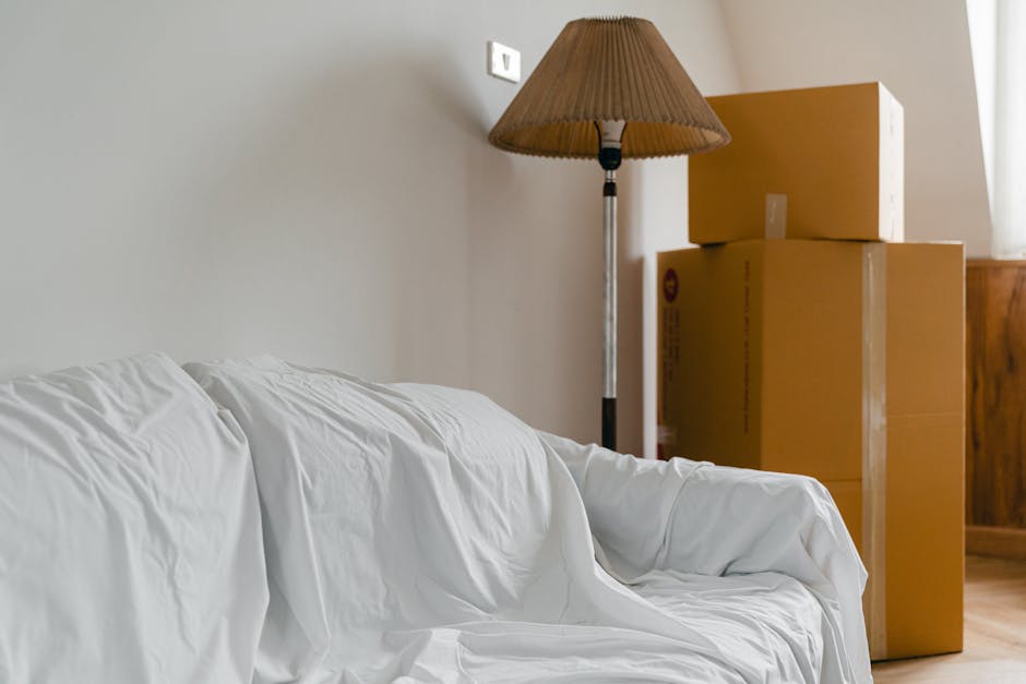 Inside a room prepared for a home relocation, a white sofa covered with a protective sheet is positioned against a light-colored wall. To the right of the sofa, there are three stacked cardboard moving boxes, with the largest at the bottom and the smallest on top, all sealed with packing tape. Behind the boxes, a wooden piece of furniture is partially visible. A tall standing lamp with a brown lampshade is situated next to the boxes, illuminating the space evenly, creating a neutral and minimalistic environment. The room features natural lighting coming from a window on the right, which is partially visible through white curtains. The setting suggests the ongoing packing and loading process associated with furniture transport or house removals, as carried out by services like Man With a Van Finchley, aligning with the focus on removals and moving logistics.