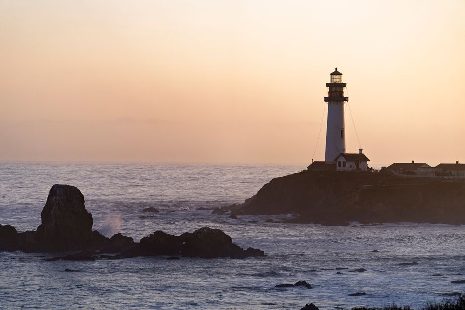 The image shows a maritime scene during sunset with a white lighthouse positioned on a rocky coastline, overlooking the ocean. The lighthouse is cylindrical with a black top and small surrounding buildings at its base. In the foreground, dark rocks protrude from the water, some with visible waves crashing against them. The scene has a calm, subdued light with a soft pastel sky, suggesting early evening or late afternoon. The ocean extends to the horizon with gentle waves, and the coastline appears rugged with uneven terrain. This setting could be associated with coastal navigation or navigation aids typical of England's seaside areas. When considering house removals or home relocation services, this coastal environment highlights the importance of careful planning for transport and logistics, especially when shifting household items near waterfront properties. Man With a Van Finchley often manages property moves in various environments, including coastal regions, ensuring secure transportation and efficient packing during the loading process, as seen in their comprehensive removals services.