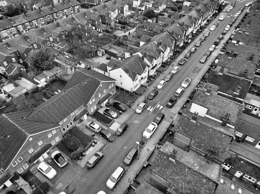 A black-and-white aerial photograph of a residential street showing closely spaced houses with pitched roofs, some with small backyards and garden sheds. Cars are parked along both sides of the narrow street, with a few on the road, and vehicles are aligned parallel to the kerb. The street is lined with a mixture of terraced and semi-detached houses, with visible driveways and small front gardens. The image captures a loading process for house removals, where a man with a van from Man With a Van Finchley may be considering the arrangement of furniture, boxed items, or packing materials within the van or on the pavement, as part of a home relocation service in tight urban streets. The environment is well-lit, with clear visibility of roof tiles, street furniture, and the arrangement of vehicles, providing an overview suitable for understanding urban-sized furniture transport and packing and moving logistics.