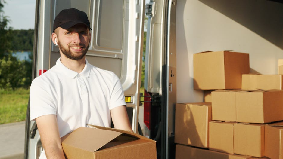 A man with a beard, wearing a white polo shirt and a black cap, stands outdoors beside the open back of a delivery van during a home relocation process. He is holding a medium-sized cardboard box, which is part of multiple boxes stacked inside the van and on the pavement. The boxes are mostly closed and made of corrugated cardboard, some wrapped with plastic film for protection. Inside the van, additional packing materials such as plastic wraps and blankets can be seen, suggesting careful handling of furniture and fragile items. The scene takes place in daylight, with a background view of greenery and trees, indicating a suburban or residential area. The loading area appears organized, with the boxes positioned near the van’s open rear doors, ready for transportation as part of a furniture transport or packing and moving operation conducted by Man With a Van Finchley, showing typical loading and lifting actions involved in house removal services near Ballards Lane, North Finchley.