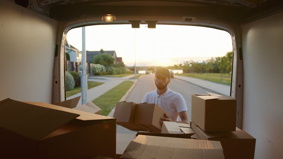 A man wearing a white polo shirt, sunglasses, and a cap stands outside at sunset, seen through the open back of a moving van. The van's interior is visible, filled with various brown cardboard boxes of different sizes, some partially open, ready for transportation. Several boxes are stacked in the van's cargo space, which appears to be clean and organized for a home relocation. The man is in the process of loading or unloading the boxes, suggesting a furniture transport or packing and moving activity as part of an upcoming house move in North Finchley N12. The background includes residential houses, a sidewalk, greenery, and a lamppost, with the evening light casting a warm glow over the scene. This image illustrates the practical steps involved in home removals, with Man With a Van Finchley delivering professional moving services, including packing, loading, and transportation in the Finchley area.