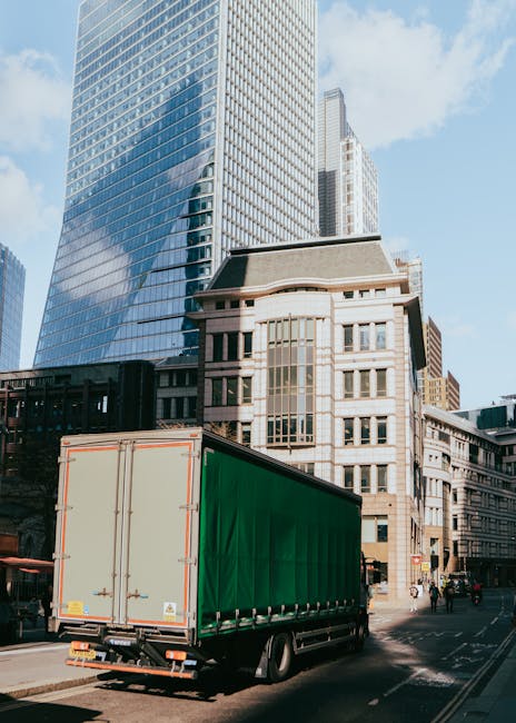 A large green cargo van parked on a city street in Finchley, with tall modern glass buildings and historical stone structures in the background. The van is positioned adjacent to the pavement, and the rear doors appear to be closed. The street is busy with a few pedestrians and vehicles, highlighting urban surroundings suitable for house and office relocations. The image captures a typical scene of furniture and household item transport during a home or commercial move, with clear daylight illuminating the scene. The presence of the van and city environment reflects professional removal services such as those offered by Man With a Van Finchley, focused on efficient furniture transport and logistical support for house removals in the area.